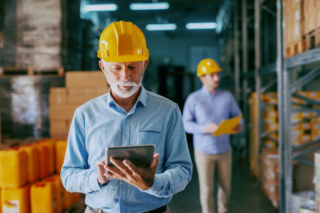 Serious Caucasian senior adult auditor in formal wear and with protective yellow helmet on head using tablet for checking on goods. In background younger manager looking at shelves and holding folder. Man on a tablet in a warehouse