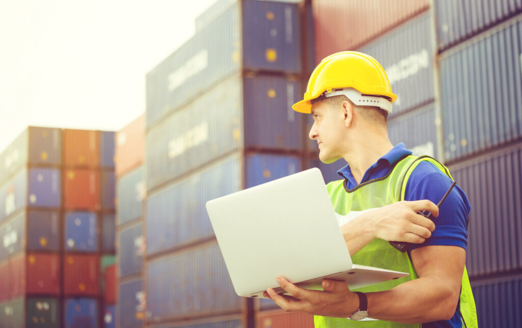 Man in a hard hat with a laptop, with shipping containers behind him