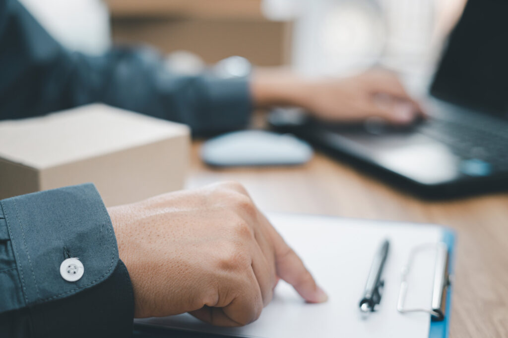 A man check Order or list inventory in stock from document and i A hand pointing at a clipboard, with a laptop in the background